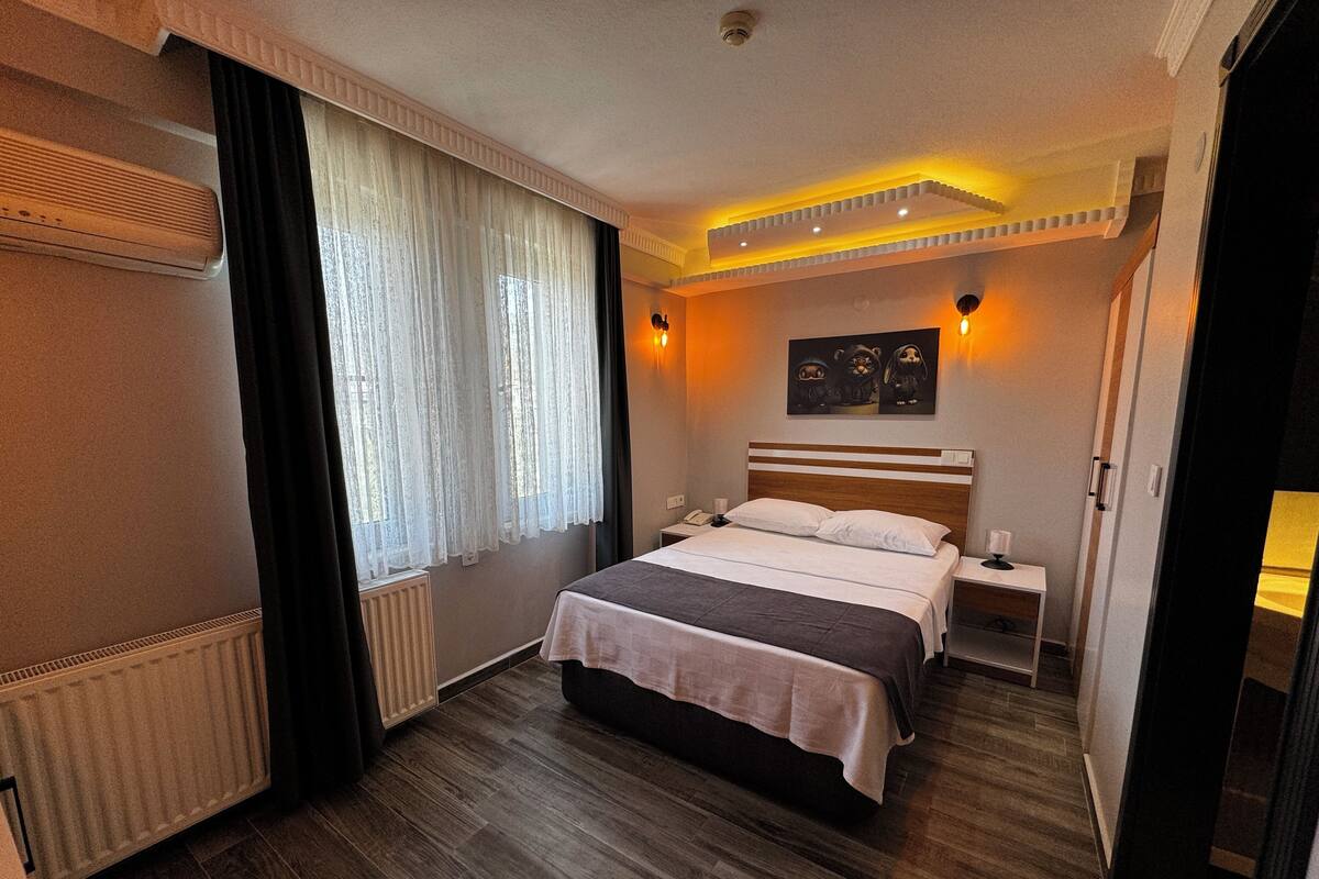 Quarto de hotel com cama, mesas de cabeceira, radiador, janela com cortinas e uma obra de arte na parede.