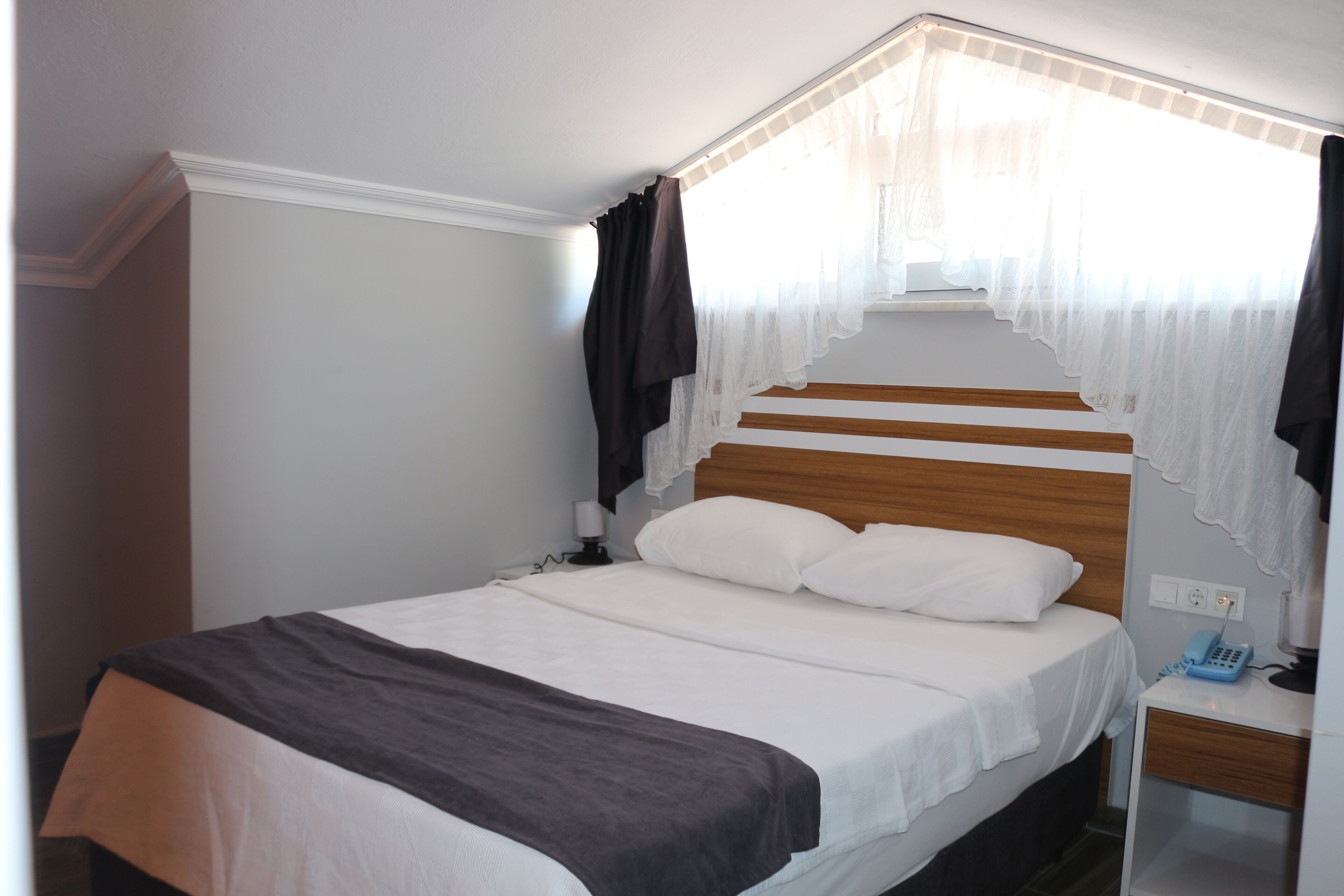 Una cama bien hecha con cabecera de madera, una mesita de noche con lámpara y una ventana con cortinas traslúcidas.