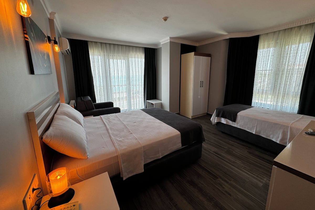 Quarto de hotel com uma cama grande, uma cama menor, uma escrivaninha, uma cadeira, um guarda-roupa e uma janela com cortinas.