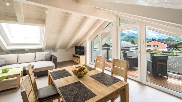 A modern interior with a dining area, a sofa, and a balcony with a view of mountains. - Villa Alpenpanorama (Ohlstadt)