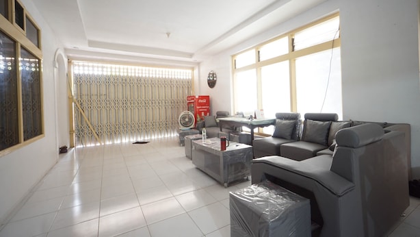 A living room with a sofa, coffee table, and a fan. There is a window with blinds and a door with a security gate. - OYO 93682 Blessing's Kediri Homestay Syariah (Kediri)