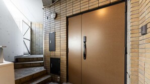 Una escalera que lleva a una puerta de madera con manija, un accesorio de luz en la pared y un pequeño panel de control.