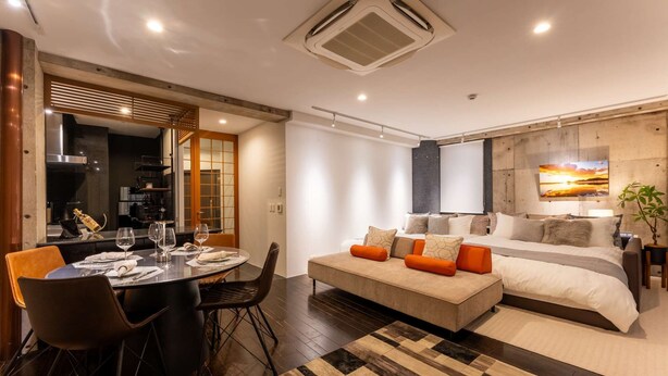 Habitación de hotel moderna con zona de comedor, sofá con cojines naranja, cama con televisor grande y suelo de madera.