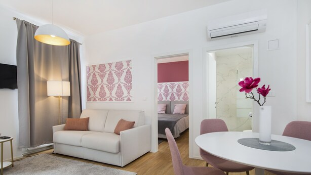 Un soggiorno moderno con un divano bianco, un tavolo da pranzo rotondo con sedie rosa, un tappeto grigio e una TV a parete.