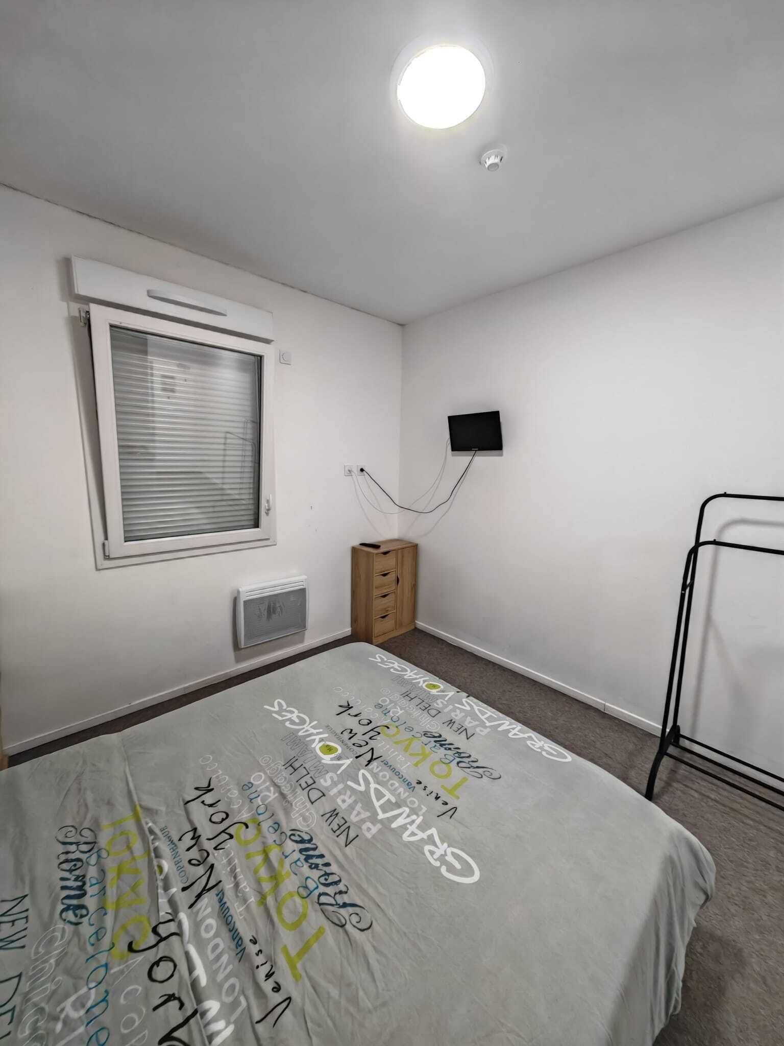 A bedroom with a bed, a window with blinds, a wall-mounted TV, a radiator, and a small wooden cabinet.