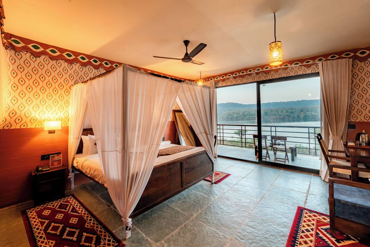 A bedroom with a bed, a ceiling fan, a nightstand, a bedside table, a window with curtains, and a view of the outdoors.