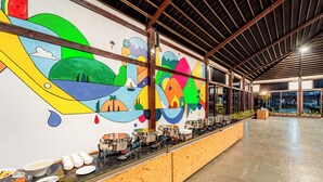 Zona de buffet con un mural llamativo, una larga mesa con diversos platos y un interior espacioso con techo alto.