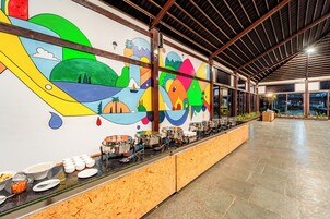 Zona de buffet con un mural llamativo, una larga mesa con diversos platos y un interior espacioso con techo alto.