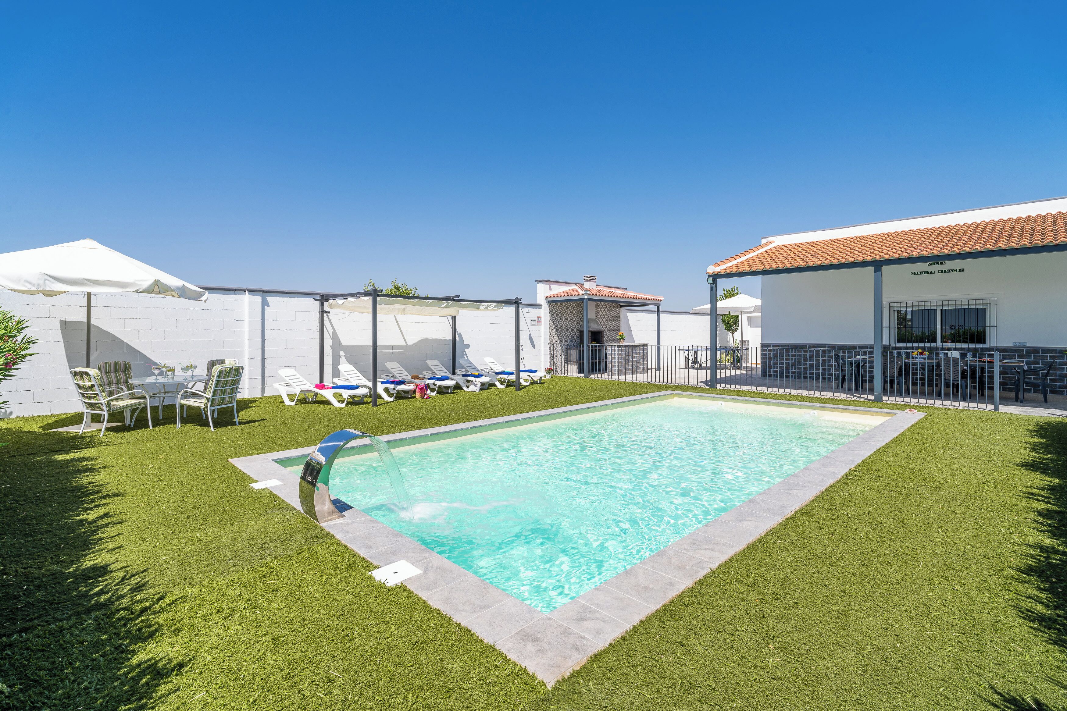 Une piscine dotée d’une terrasse carrelée, entourée d’une clôture blanche et agréée d’un espace de repos couvert avec des chaises et des parasols.