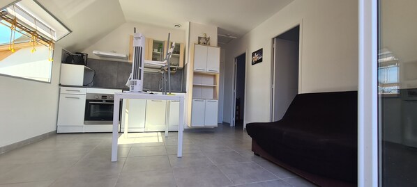 A modern kitchen with white cabinets, a built-in oven, and a central island. There is a black sofa in the living area and a glass door leading to another room. - Tipi des Dunes a 800m de la Plage. Les Animaux de Compagnie Sont les Bienvenues (Surtainville)