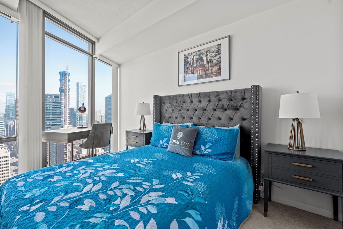 Habitación de hotel con una cama grande, vista a la ciudad, un cuadro en la pared y un escritorio con una lámpara.