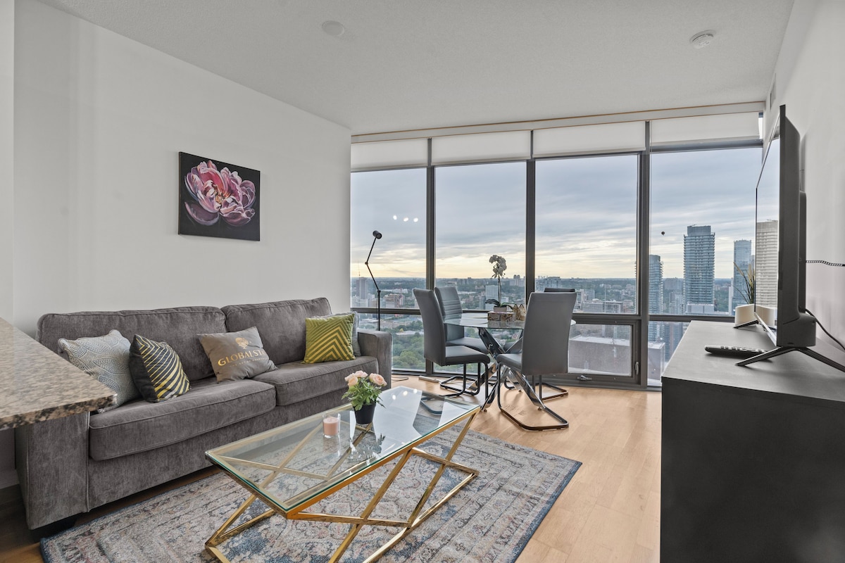 A modern living room with a grey sofa, a glass coffee table, and a view of the cityscape through large windows.