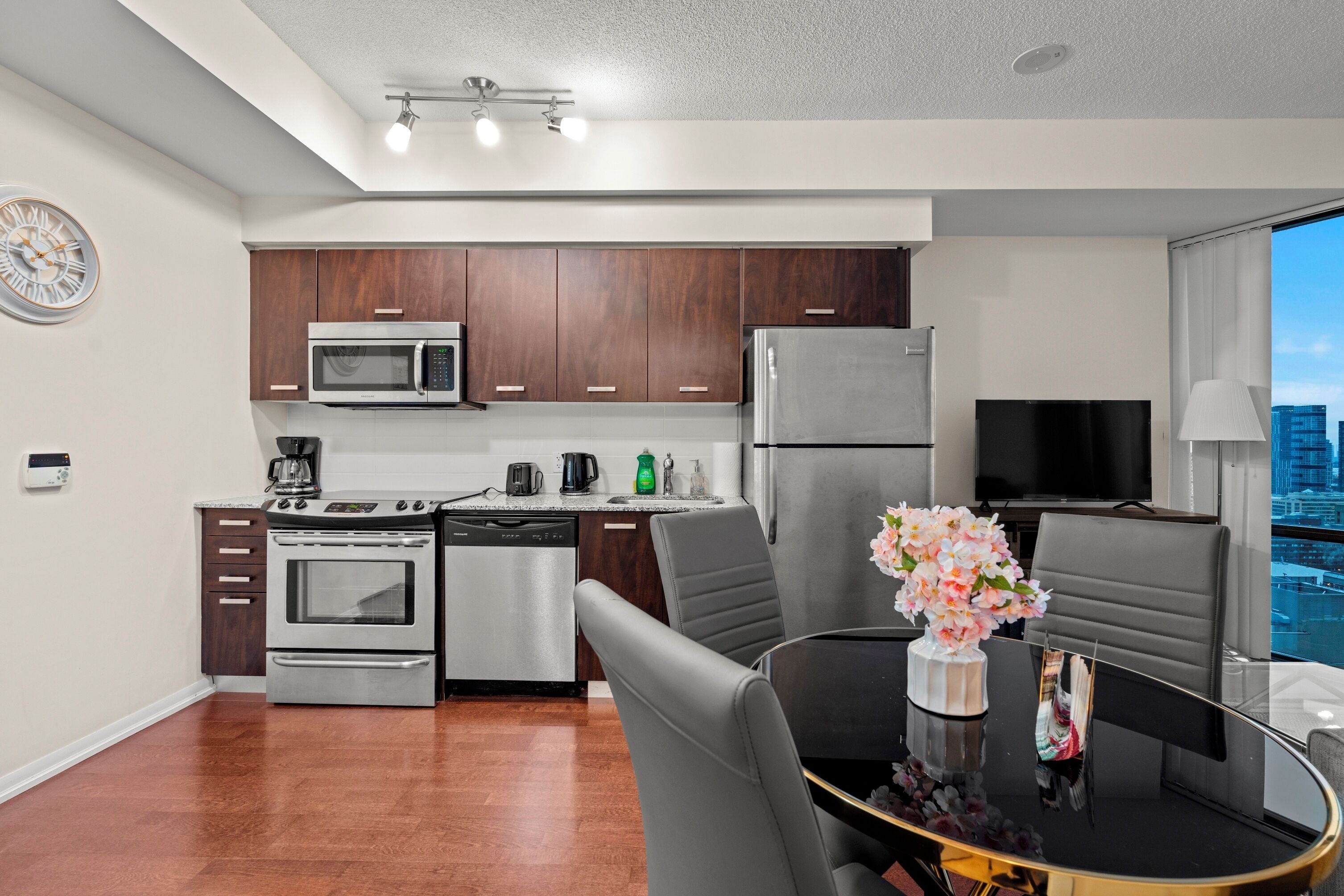 A modern kitchen with stainless steel appliances, a dining area with a glass table, and a city view through large windows.