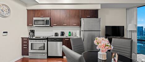 A modern kitchen with stainless steel appliances, a dining area with a glass table, and a city view through large windows.