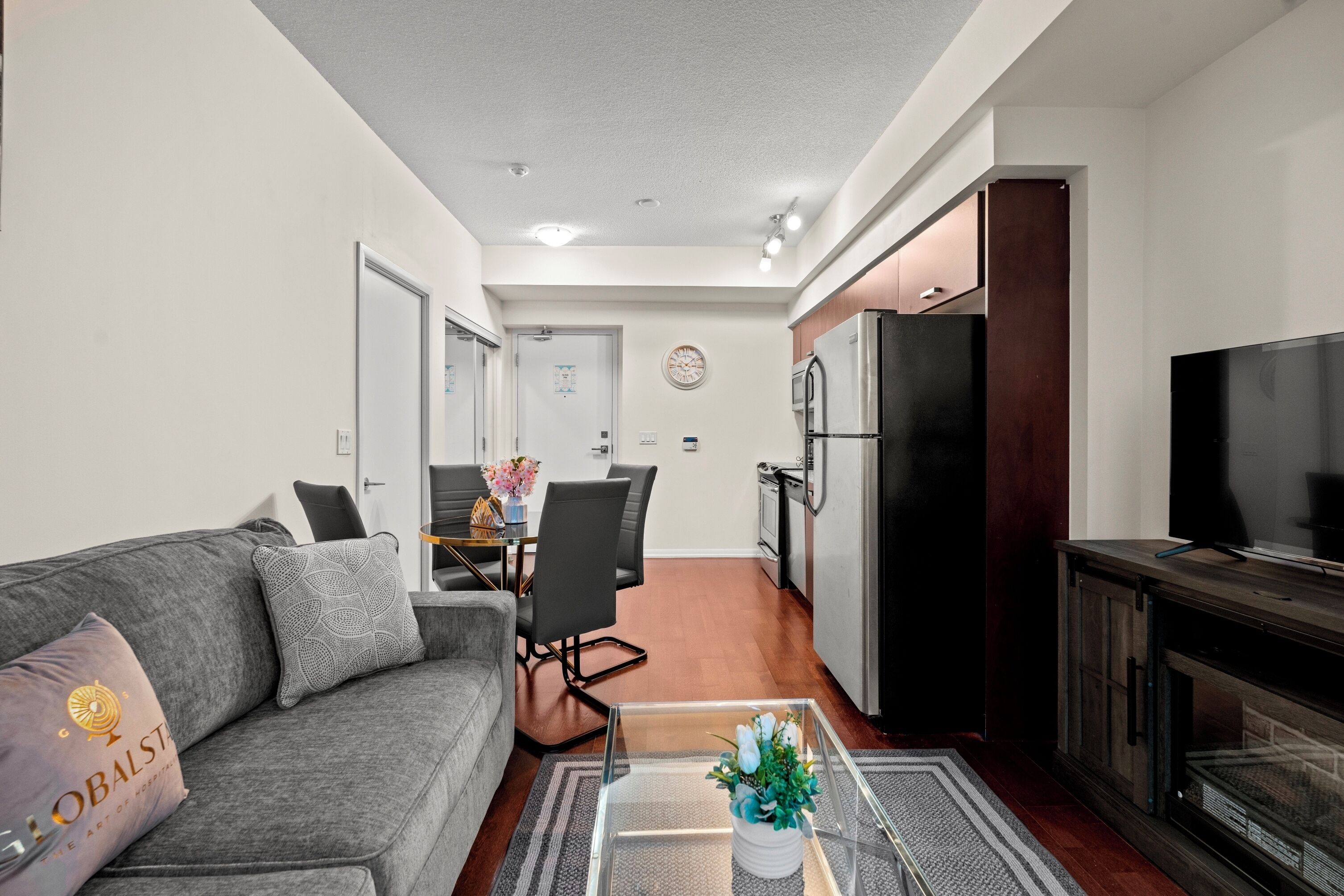 Ein modernes Wohnzimmer mit grauem Sofa, einem Couchtisch und einem Fernseher. Es gibt einen Essbereich mit Tisch und Stühlen sowie eine Küche mit Kühlschrank und Herd.