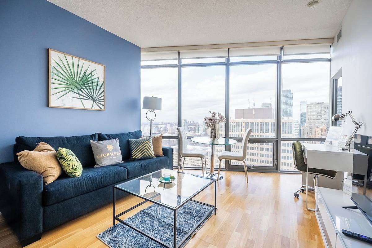 Un salón moderno con un sofá azul oscuro, una mesa de centro de vidrio y vistas a la ciudad a través de amplios ventanales.