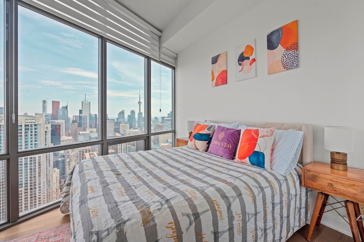 Un dormitorio con un ventanal grande, una cama con ropa de cama a rayas, almohadas coloridas y arte abstracto en la pared.