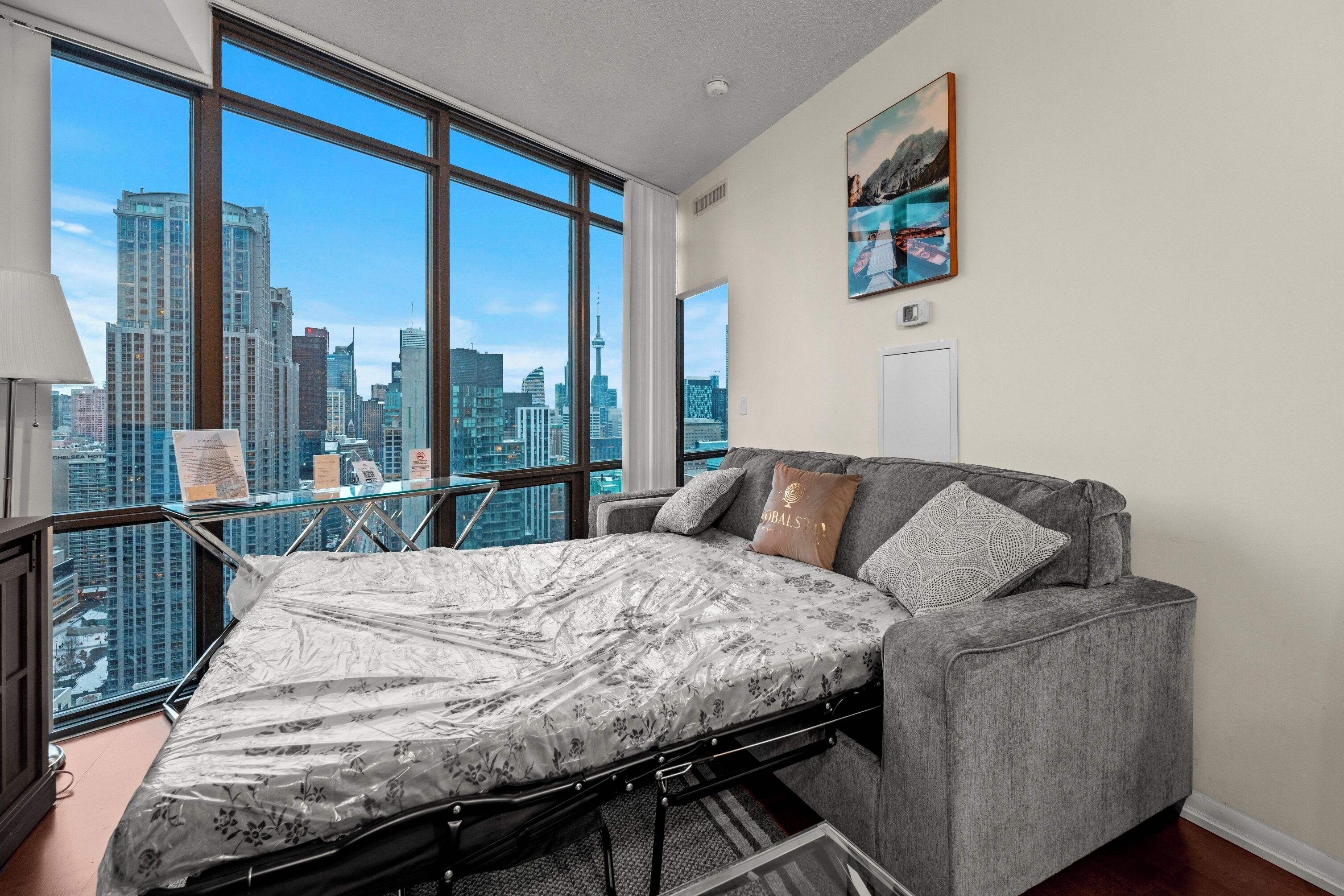 A modern bedroom with a convertible bed, large windows with a city view, a balcony, and a painting on the wall.