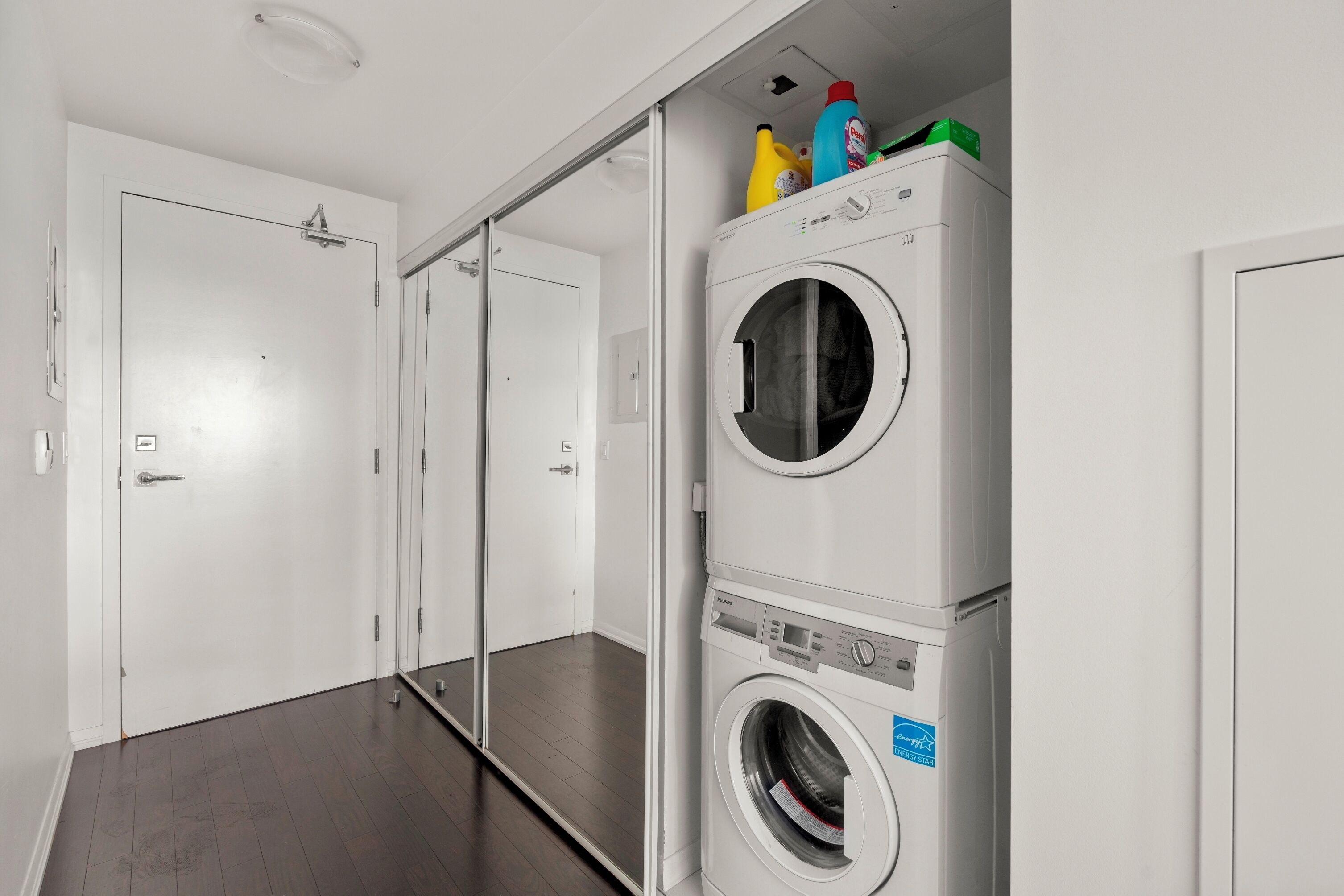 Un lavadero con lavadora y secadora de carga frontal, un espejo y una puerta que da a otra habitación.