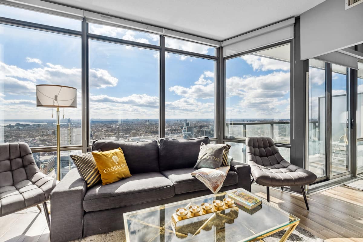 Una sala moderna con un sofá gris, dos sillones, una mesa de centro de vidrio y una lámpara de pie. Amplias ventanas ofrecen vistas a la ciudad.