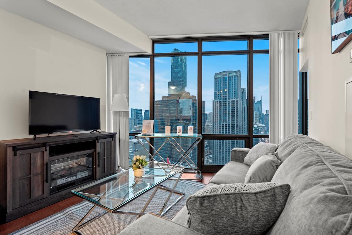 Una sala de estar moderna con un televisor grande, una mesa de centro de vidrio, un sofá y vistas a un horizonte urbano a través de amplios ventanales.