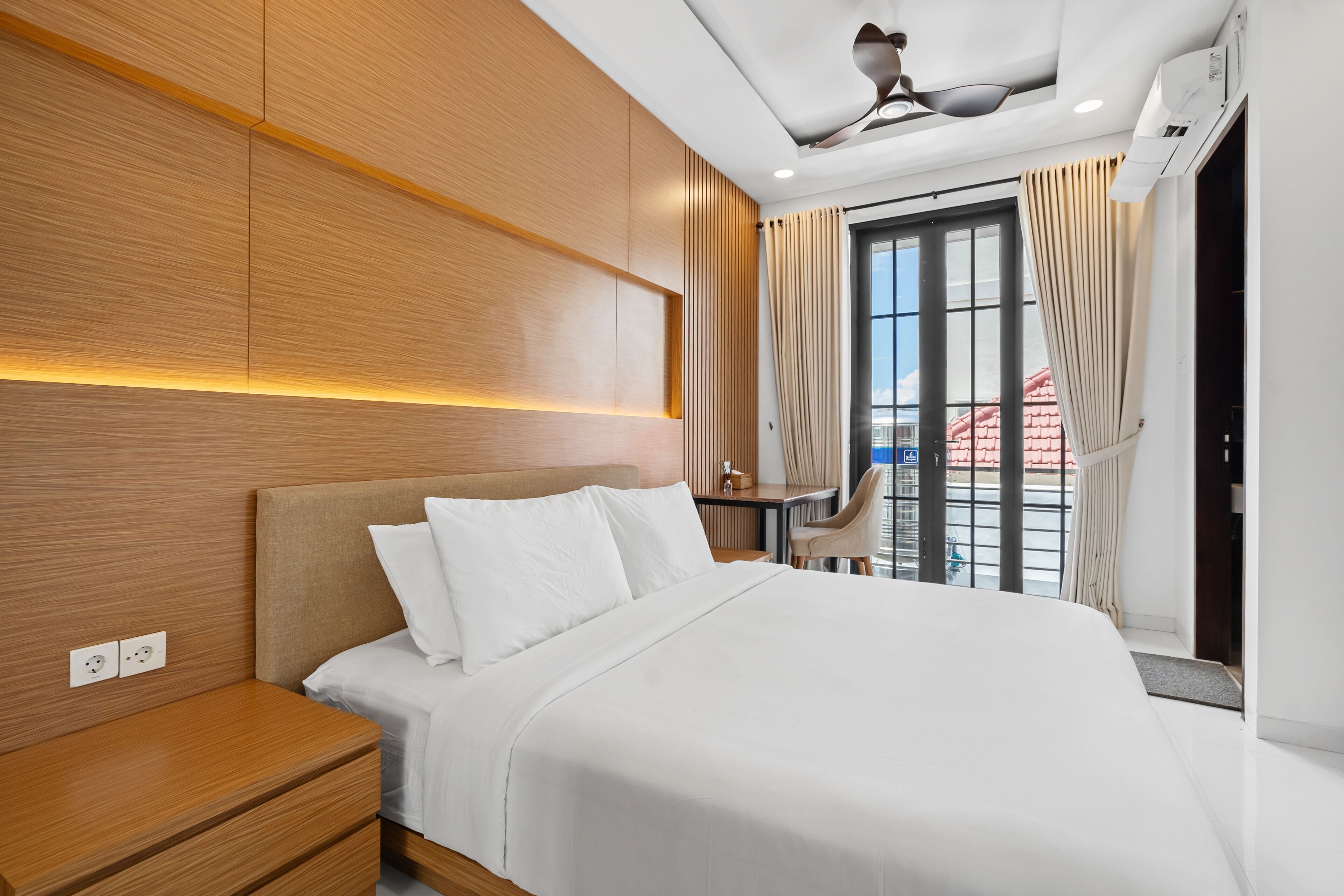 A modern hotel room with a large bed, wooden panel walls, a ceiling fan, and a view of the city through a large window.