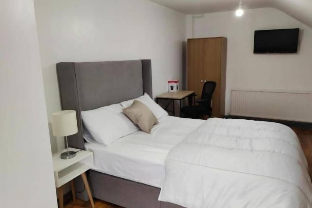 Ein Schlafzimmer mit einem Bett, einem Nachttisch mit Lampe, einem Sessel, einem kleinen Tisch und einem an der Wand befestigten Fernseher.