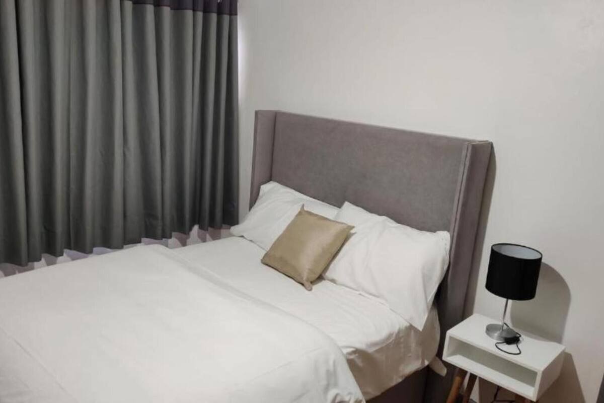 Ein ordentlich bezogenes Bett mit weißer Bettwäsche und einem einzelnen beigen Kissen, ein grauer möbelbezogener Kopfteil, ein kleiner Nachttisch mit einer schwarzen Lampe und graue Vorhänge.