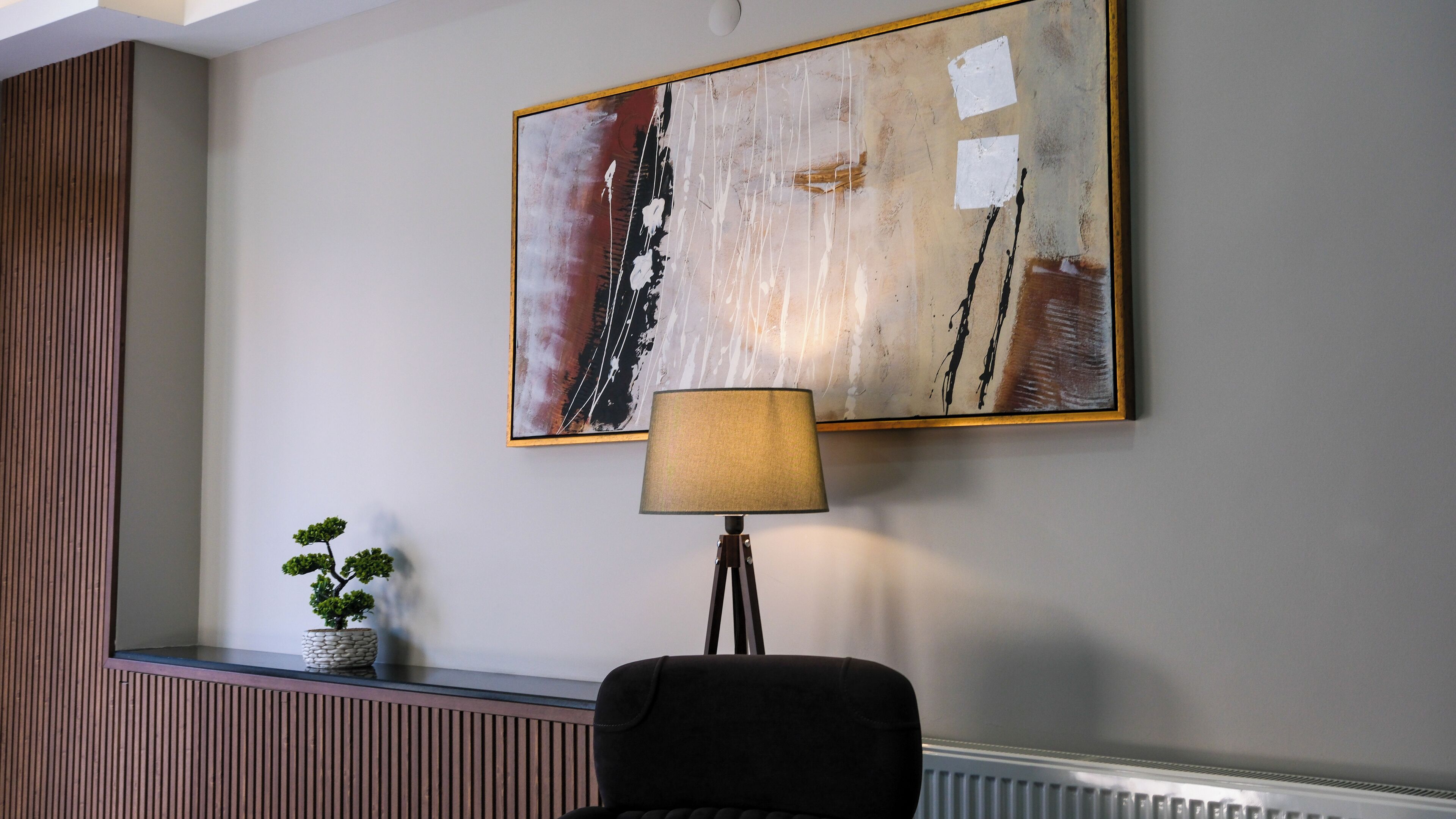 A modern room with a minimalist design, featuring a wall-mounted abstract painting, a floor lamp, a potted plant, and a black chair.