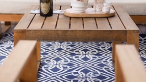 A wooden table with a bottle of wine, a glass, a decorative bowl with flowers, and small jars on a patterned rug.