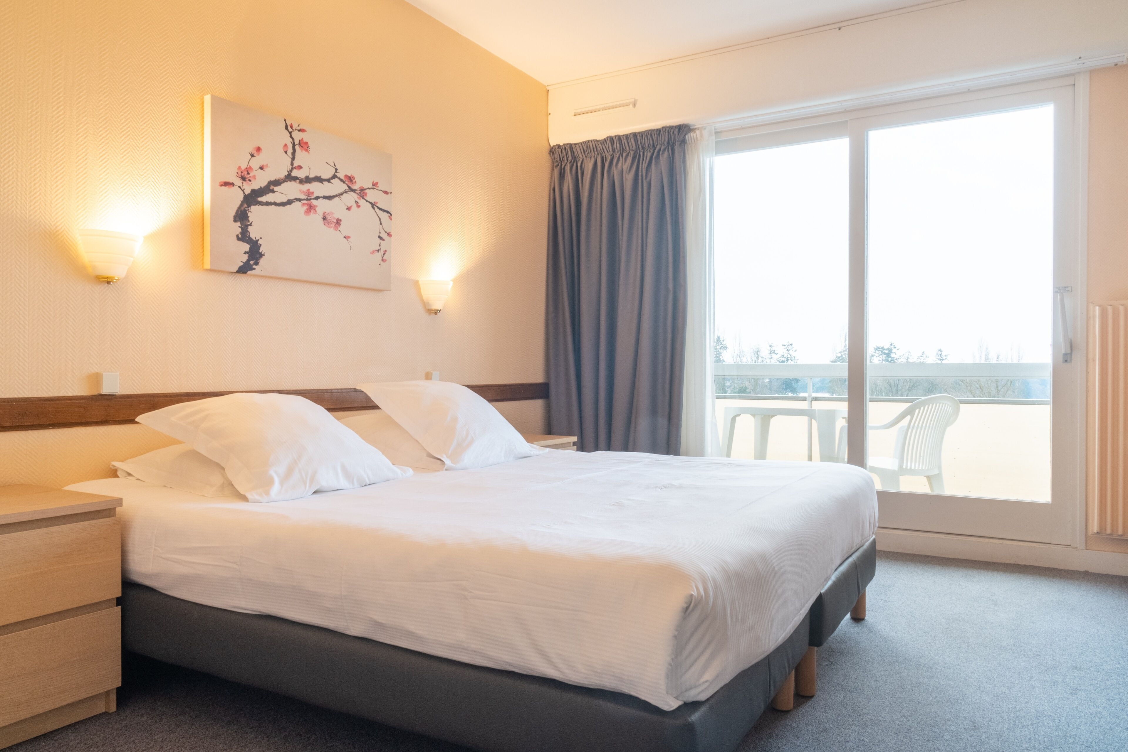 A hotel room with a bed, a nightstand, a wall-mounted lamp, a window with curtains, and a balcony view.