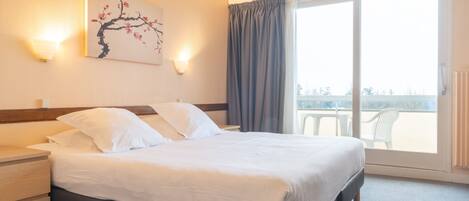A hotel room with a bed, a nightstand, a wall-mounted lamp, a window with curtains, and a balcony view.