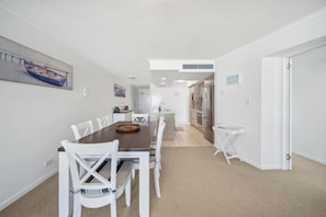 A dining area with a long table, white chairs, a painting of a boat, and a kitchen in the background.