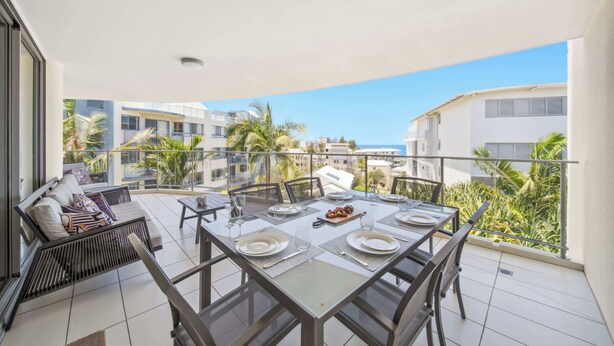 A balcony with a dining table set for four, a sofa, and a view of other buildings and palm trees. - Seaside Serenity: Apartment with Ocean Views (Kings Beach)