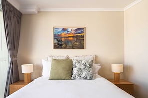 A neatly made bed with two bedside lamps, a painting of a sunset over a body of water, and a window with curtains.