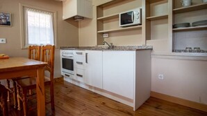 Een kleine keuken met witte kastjes, een magnetron en een spoelbak. Er staat een houten eettafel met stoelen.