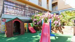 Un área de juegos infantiles con un tobogán, una casita pequeña y un parterre con flores rosadas.