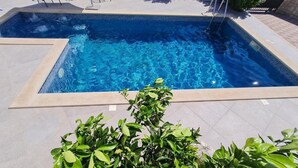 A rectangular swimming pool with clear blue water, surrounded by a tiled deck and a small green bush in the foreground.