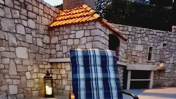 A lounge chair with a striped cushion and a pillow with the word "relax" on it, placed on a patio with a stone wall and a tiled roof in the background.
