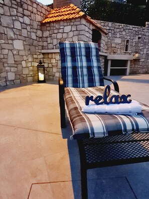 A lounge chair with a striped cushion and a pillow with the word "relax" on it, placed on a patio with a stone wall and a tiled roof in the background. - Luxury Apartment Ani (Sutivan)