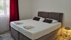 A neatly made bed with white linens and two dark pillows, a bedside lamp, and a window with red curtains.