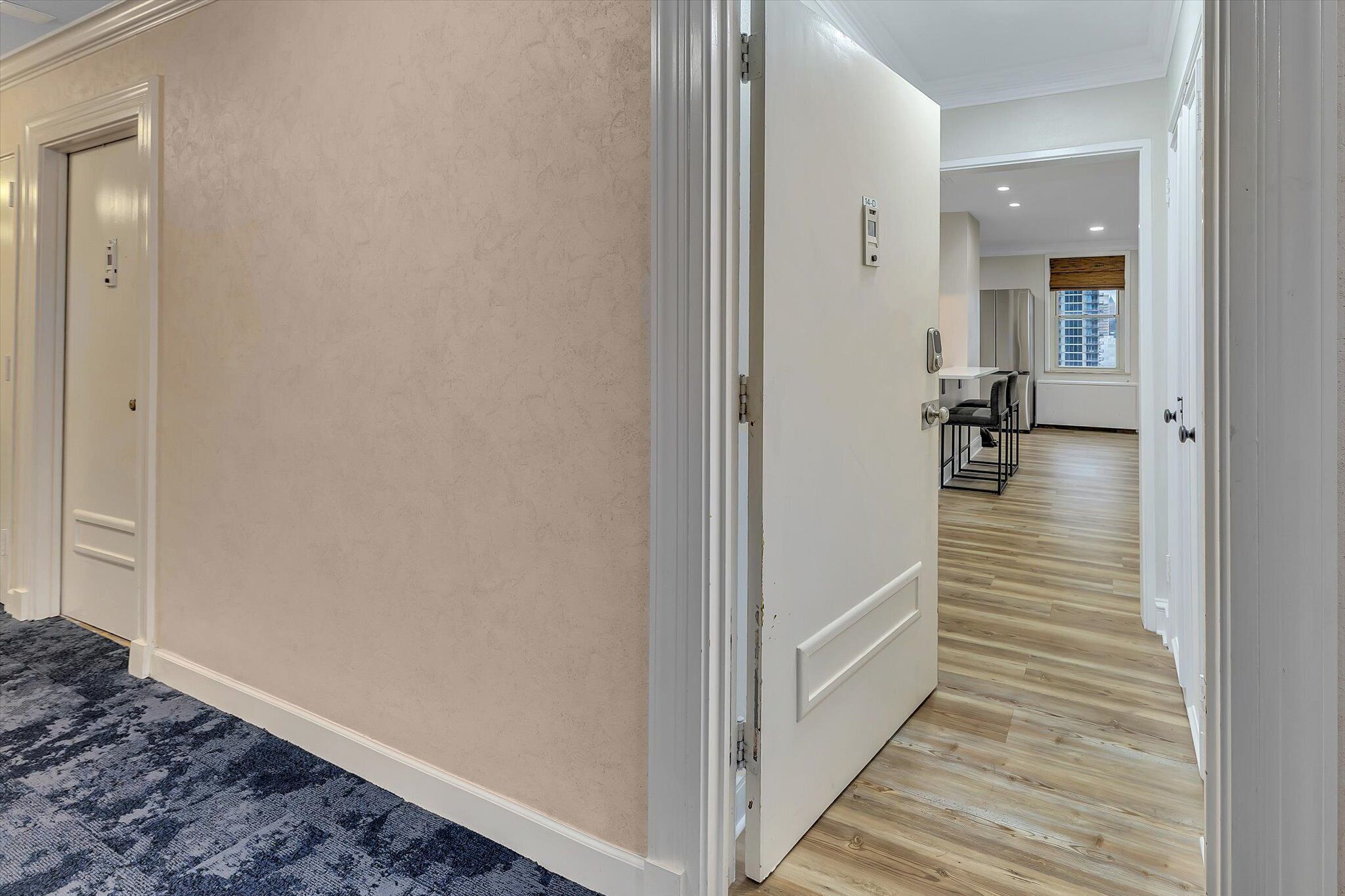 A hotel hallway with beige walls, a wooden floor, and a door leading to a room with a window.