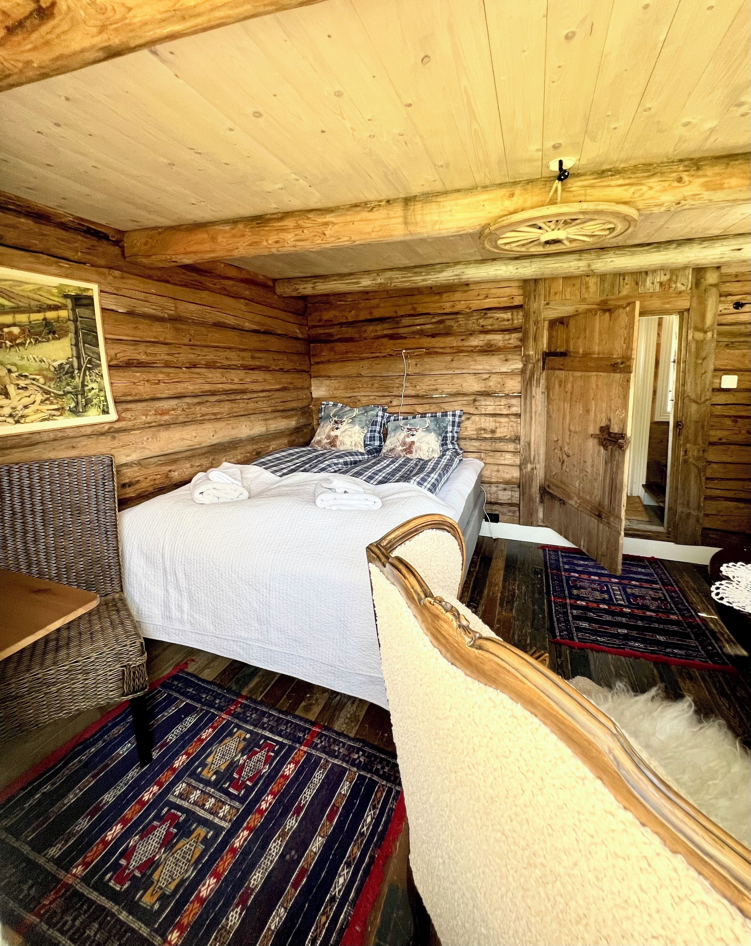 A rustic wooden cabin bedroom with a bed, a wicker chair, a patterned rug, and a wooden door.