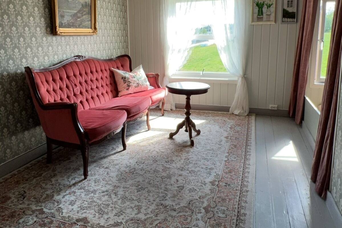 A room with a red sofa, a small round table, patterned wallpaper, and a window with curtains.