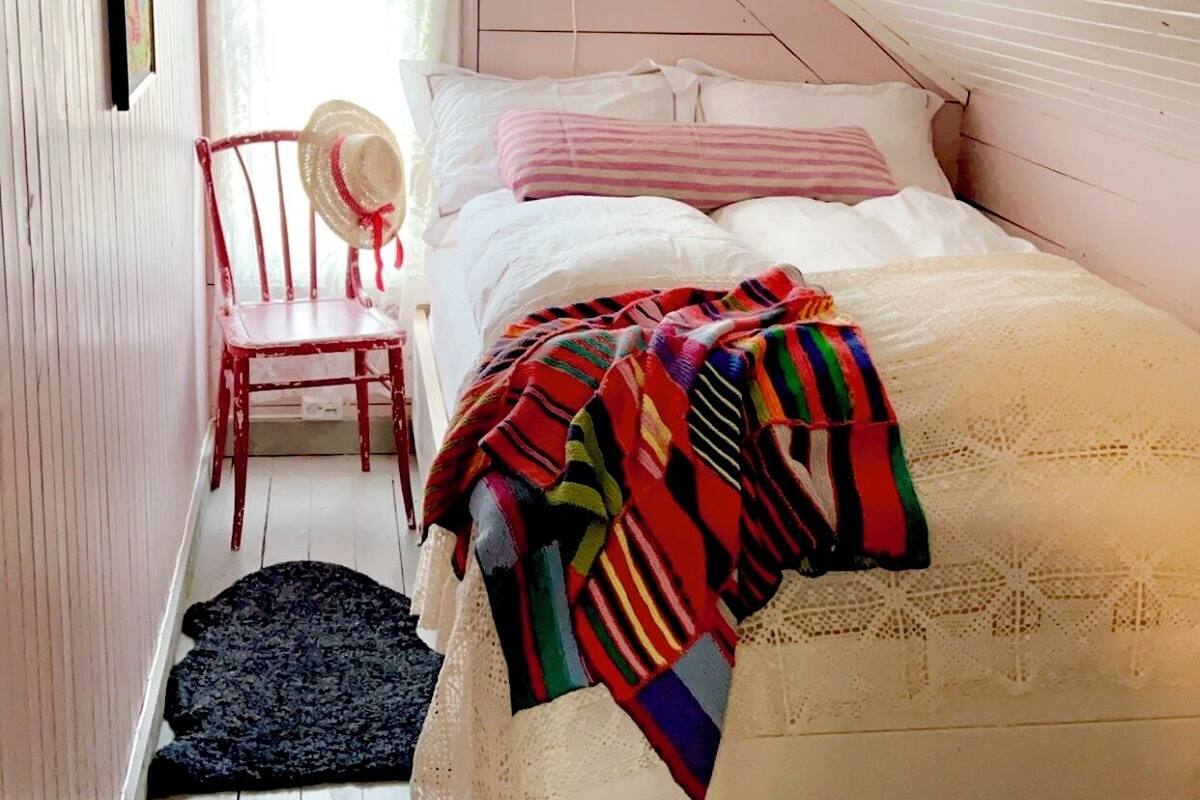 A cozy attic bedroom with a sloped ceiling, a bed with a colorful striped blanket, a chair with a hat, and a small window with curtains.