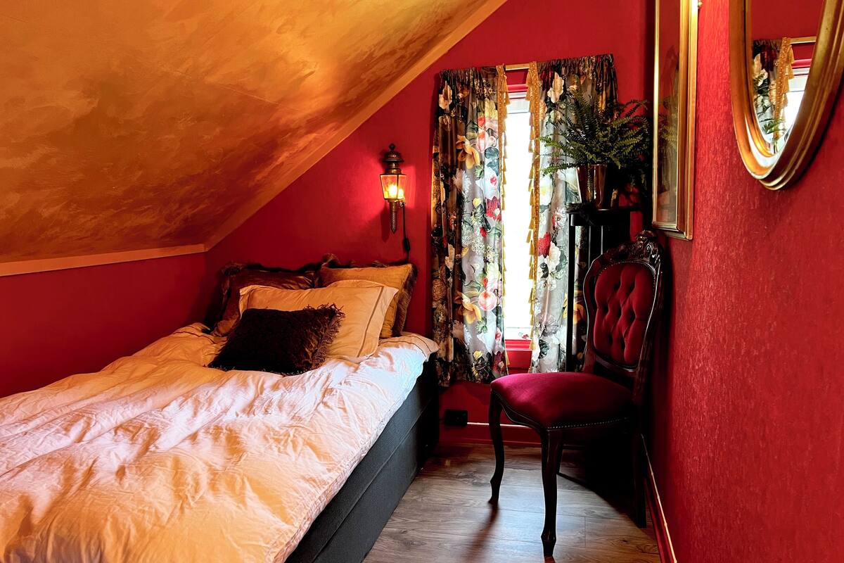 A bedroom with a sloped ceiling, a bed with white bedding, a red chair, and floral curtains.