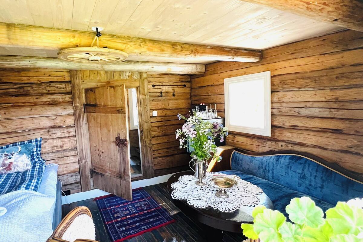 A cozy cabin interior with a wooden ceiling, a blue upholstered bench, a small table with a lace doily, and a potted plant.