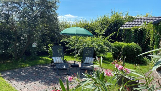 Ein Garten mit zwei Liegestühlen unter einem grünen Sonnenschirm, umgeben von üppiger Bepflanzung und blühenden Pflanzen.