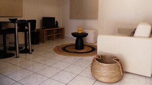 Una sala de estar con un sofá blanco, un televisor sobre un mueble de madera, una mesita negra con un florero y una cesta de mimbre.