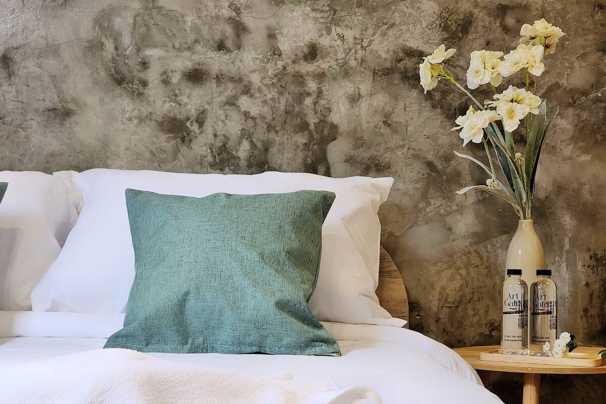 Un lit avec des oreillers blancs et turquoise, un vase rempli de fleurs blanches et deux bouteilles étiquetées « Art Gates » sur une table d’appoint en bois.